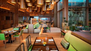 Um restaurante espaçoso com pé direito alto, luminárias pendentes e uma área central de estar com bancos estofados em verde.