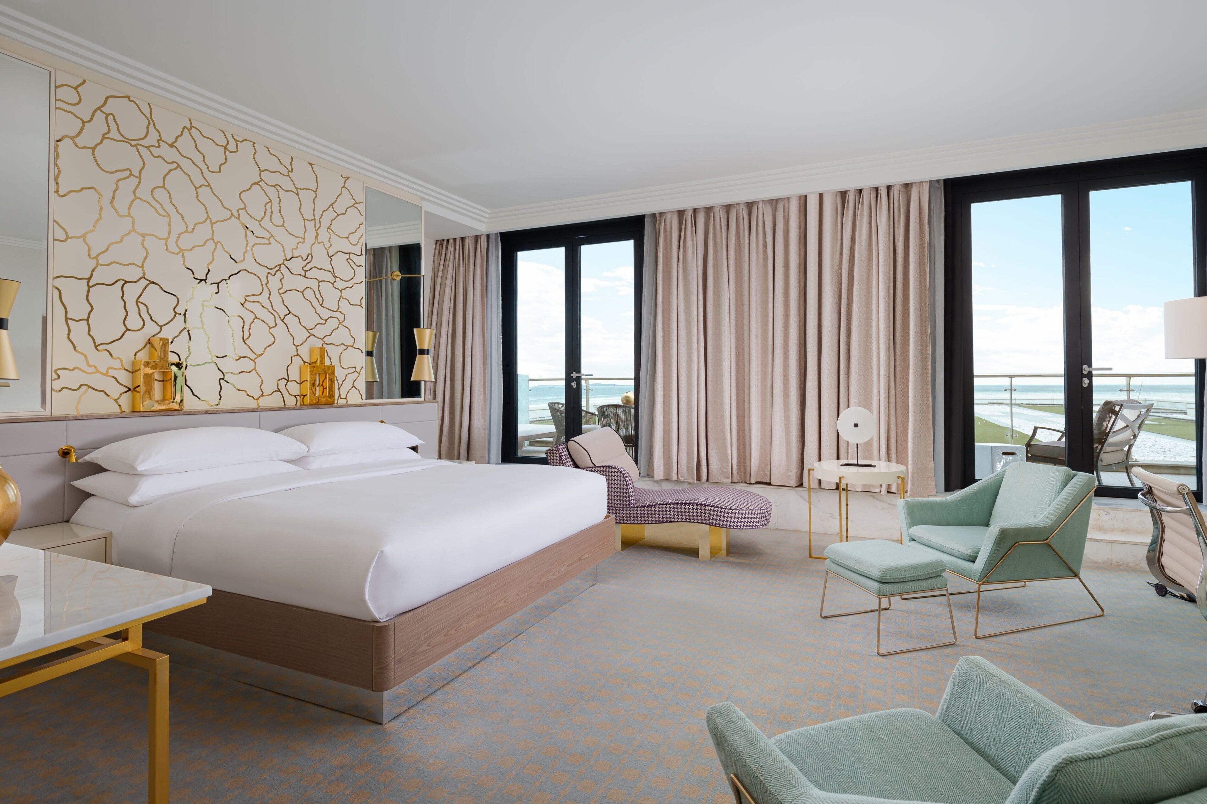 A hotel room with a large bed, a seating area with chairs, a small table, and a view of the ocean through large windows.