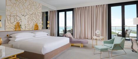 Habitación de hotel con una cama grande, zona de estar con sillas, una mesita y vista al océano a través de amplias ventanas.