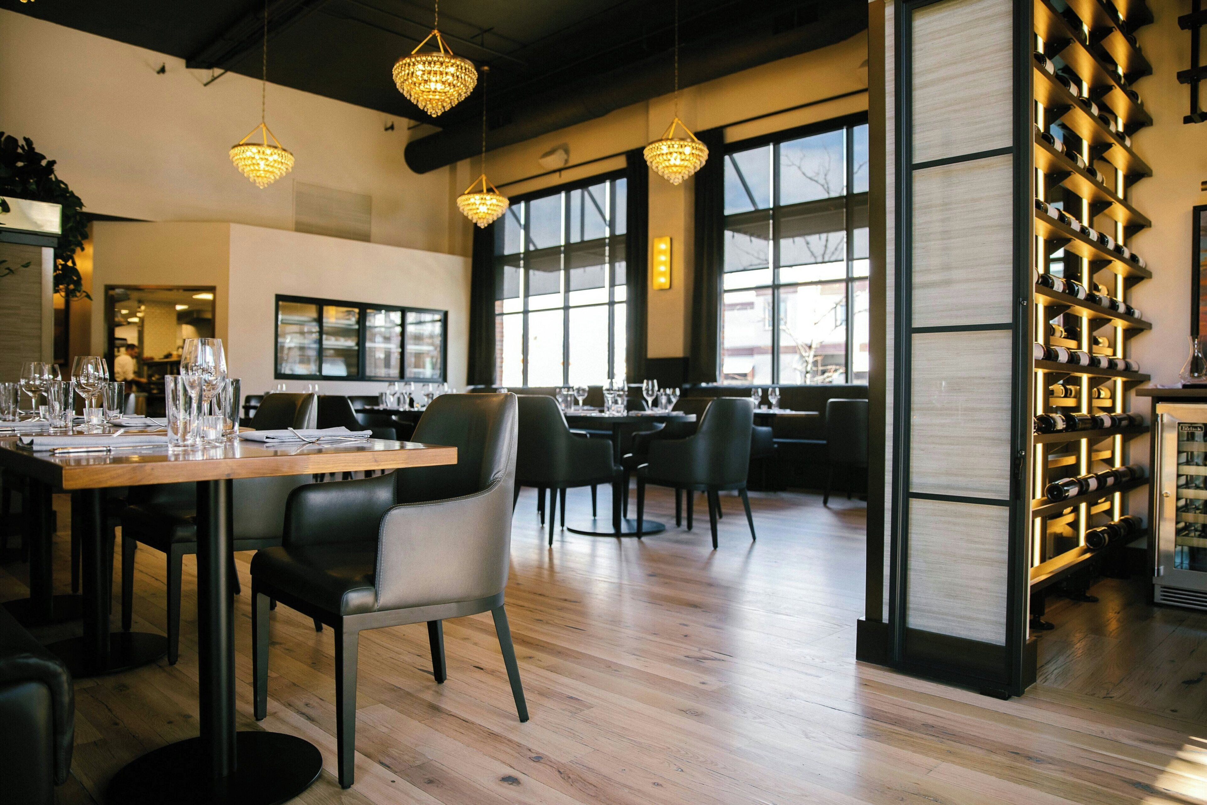 Een restaurantinterieur met houten tafels, zwarte stoelen, hangende kroonluchters en een wijnrek.