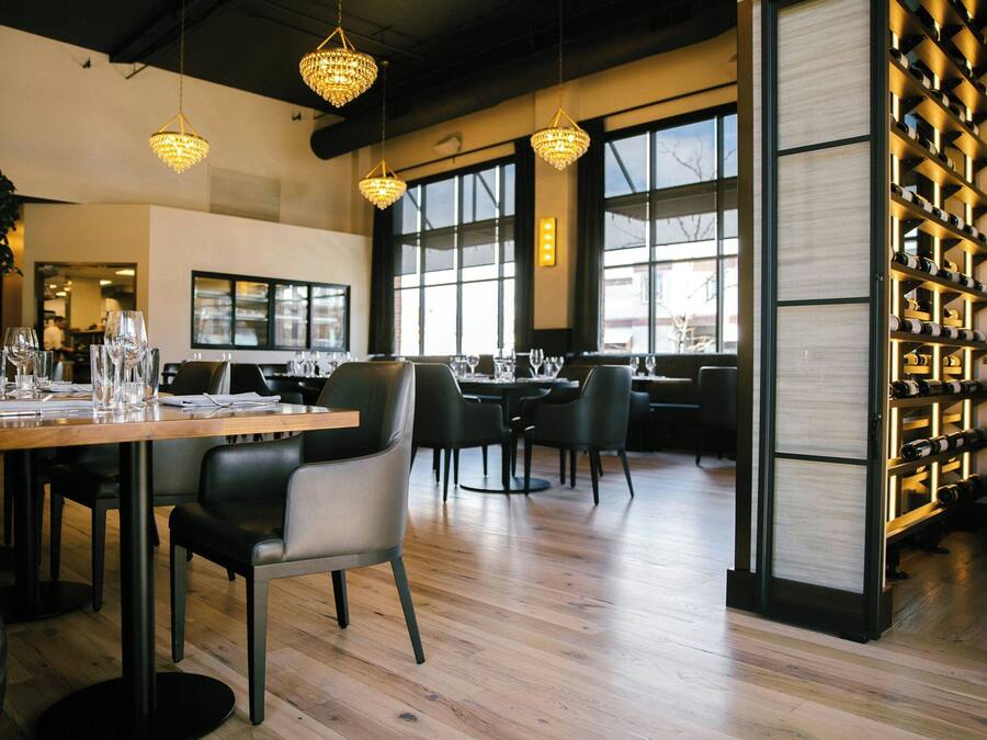 Een restaurantinterieur met houten tafels, zwarte stoelen, hangende kroonluchters en een wijnrek.