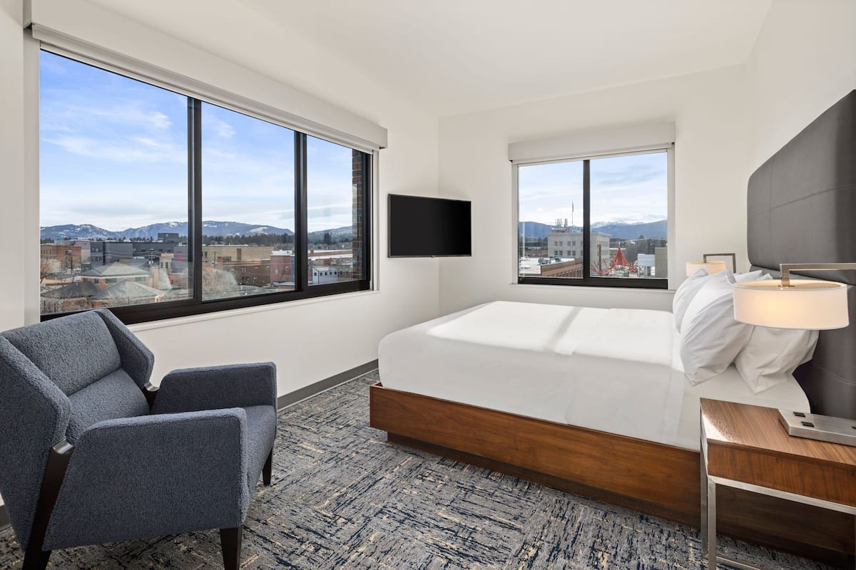A hotel room with a large bed, a TV mounted on the wall, a chair, and a view of the city and mountains.