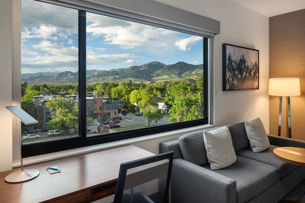A modern hotel room with a large window offering a view of mountains and a town. There is a gray sofa with pillows, a desk with a lamp, and a framed picture on the wall.