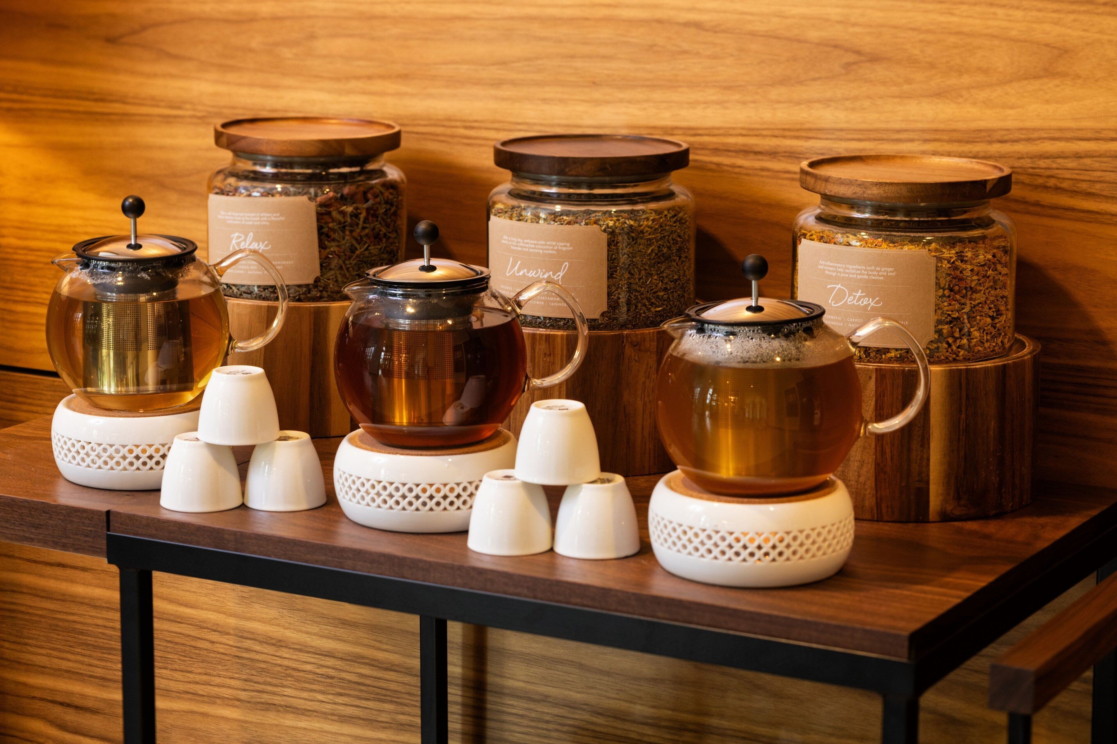 Una mesa de madera con varios dispensadores de té y café, y tazas de cerámica blanca.