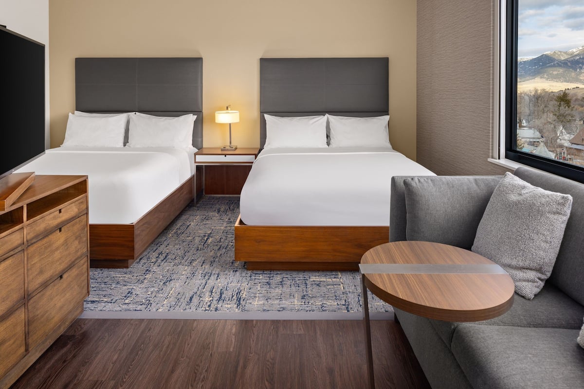 A hotel room with two beds, a wooden dresser, a gray sofa, a round table, and a view of mountains through a large window.