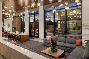 A modern hotel lobby with a central island dining area, a sofa with cushions, and a coffee table with plants.