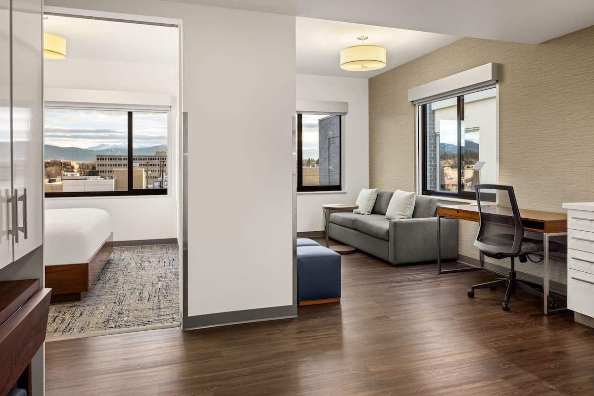 A modern hotel room with a bed, a sofa, a desk, and a large window offering a city view.