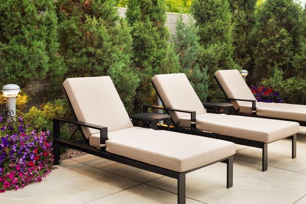 Three lounge chairs with beige cushions and black metal frames, arranged on a tiled patio with a backdrop of dense green foliage and colorful flowers.