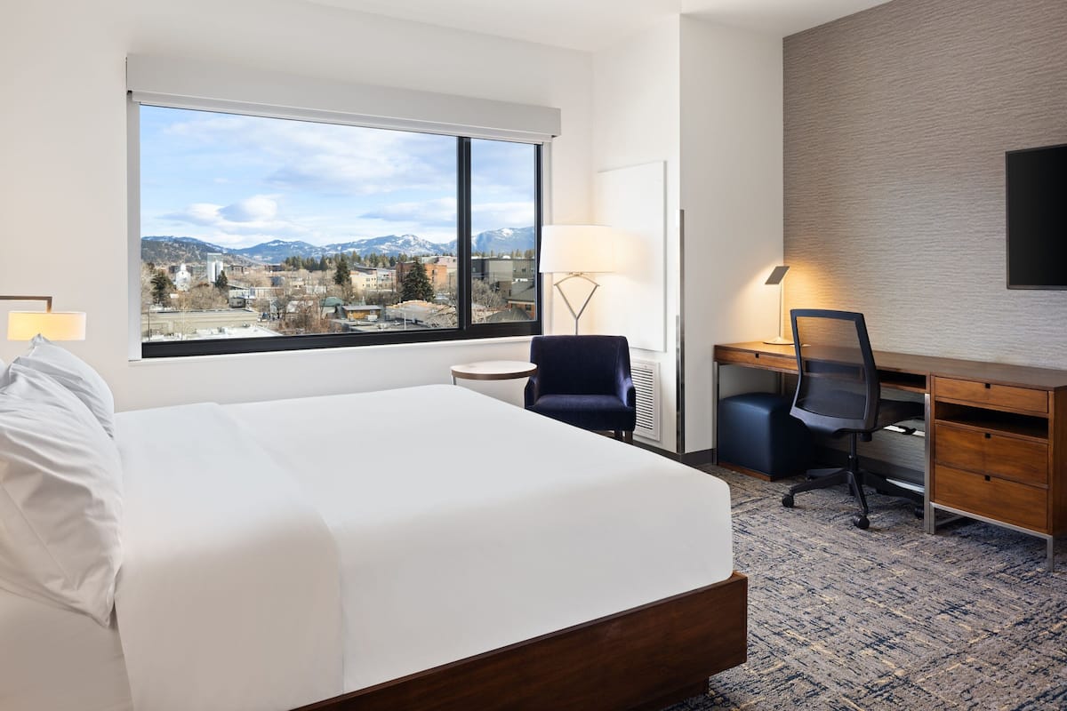A hotel room with a large bed, a desk, a chair, a lamp, and a view of a cityscape with mountains.
