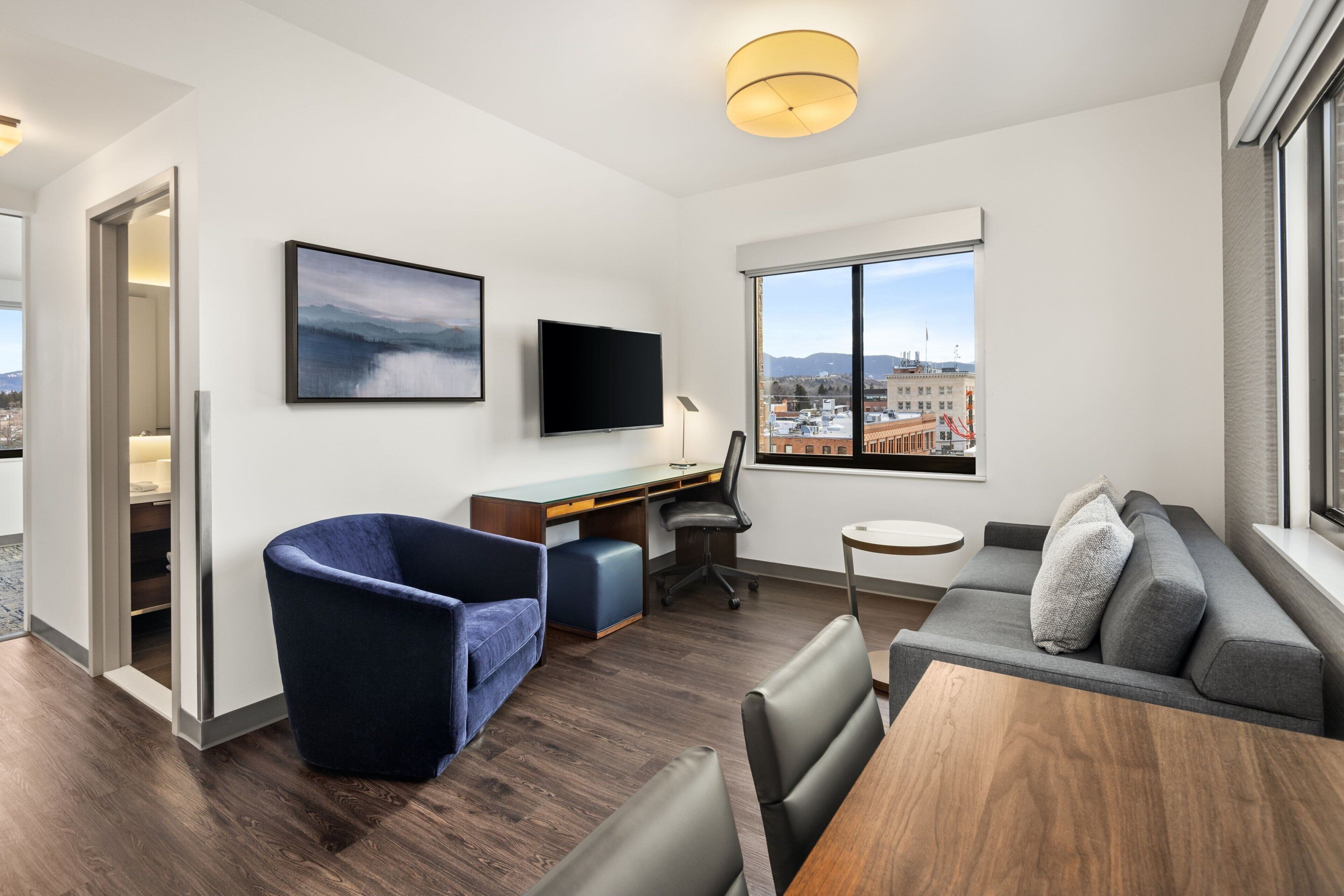 A modern living room with a desk, a blue armchair, a gray sofa, and a large window offering a city view.