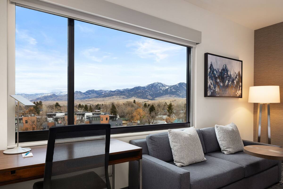 A modern hotel room with a large window offering a mountain view, a gray sofa with pillows, a desk with a chair, and a lamp.