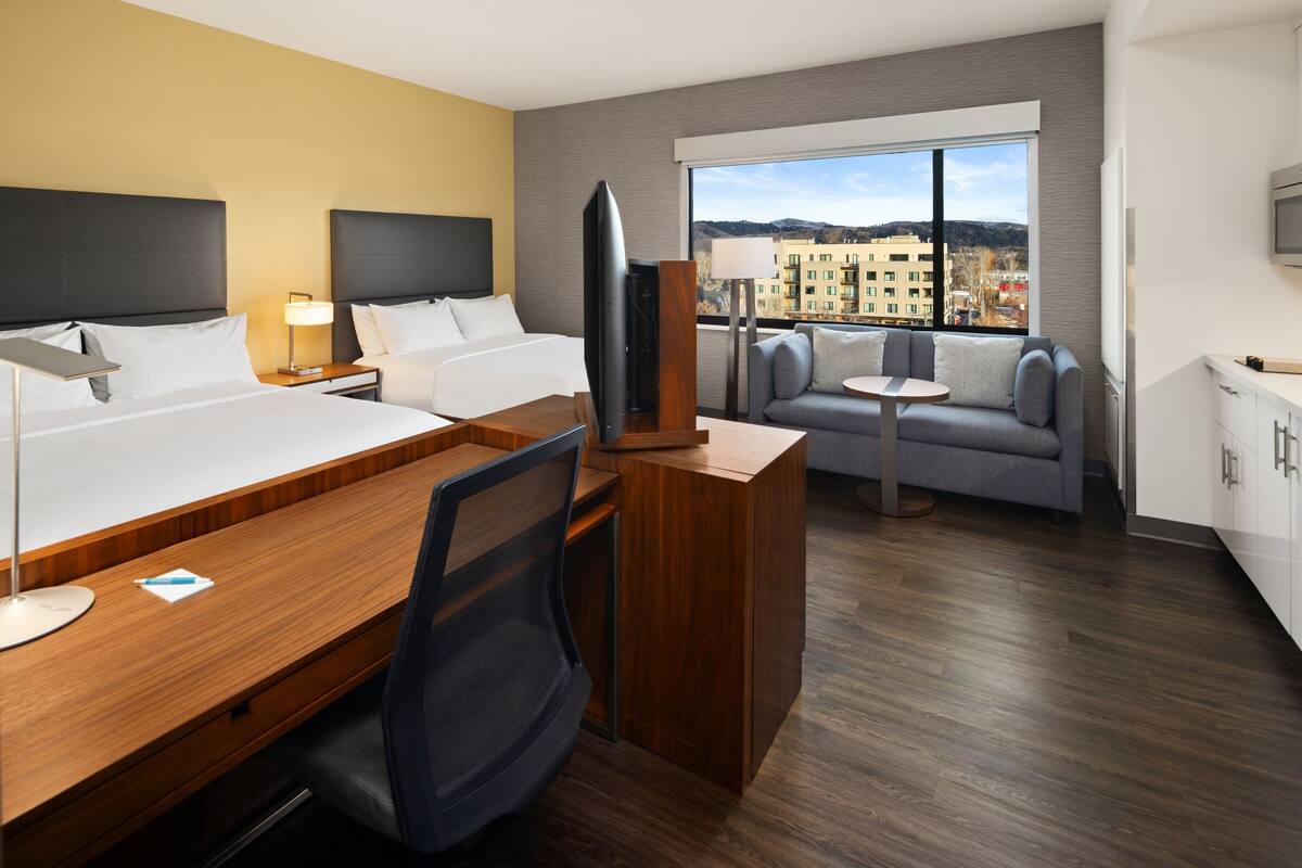 A hotel room with a bed, desk, TV, sofa, and a view of the city.