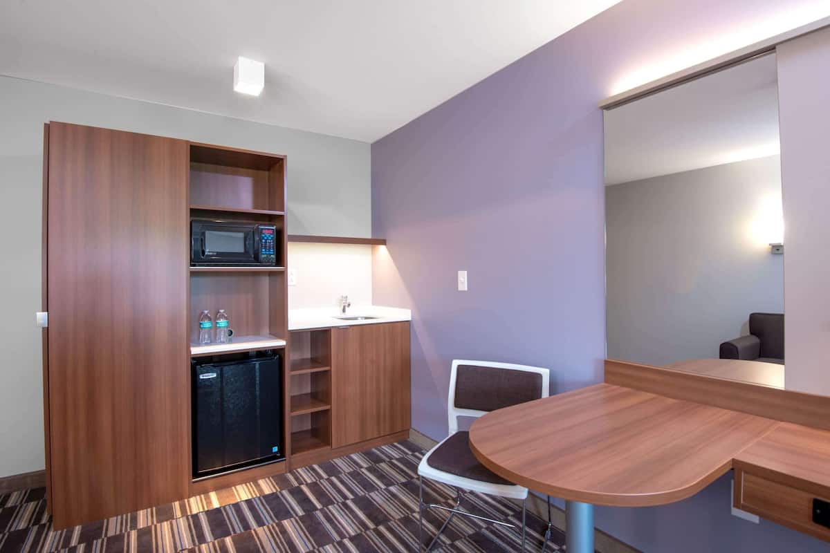 A compact hotel room with a kitchenette, a microwave, a sink, a small refrigerator, a chair, and a mirror.