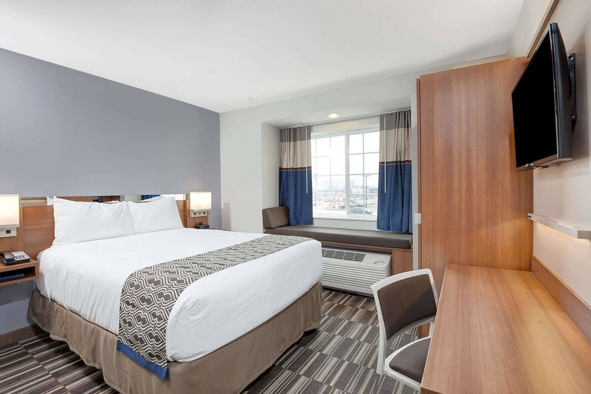 A hotel room with a large bed, bedside tables, a desk, a chair, a TV, and a window with curtains.