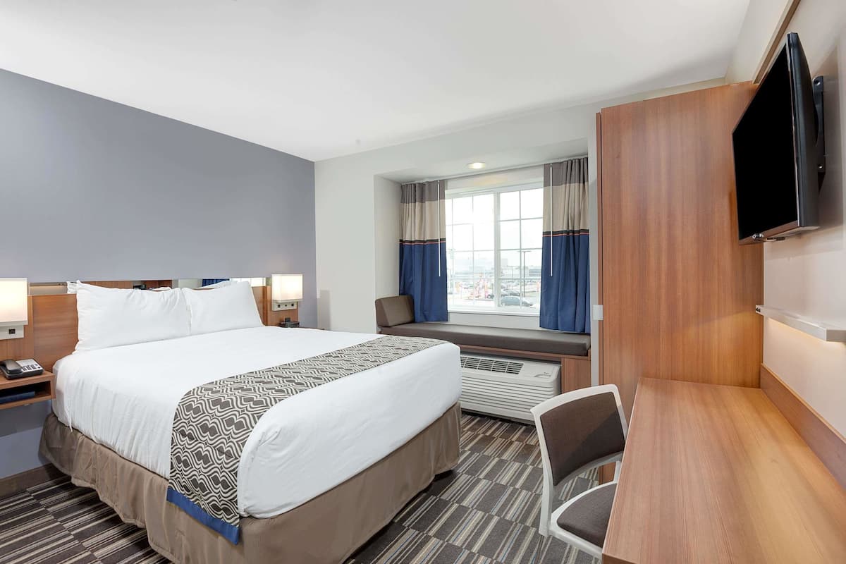 A hotel room with a large bed, bedside tables, a desk, a chair, a TV mounted on the wall, and a window with curtains.