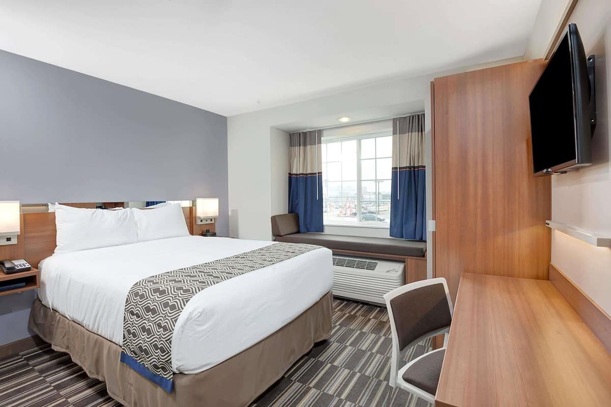 A hotel room with a large bed, bedside tables, a desk, a chair, a TV, and a window with curtains.