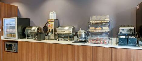 A self-service breakfast station with a microwave, toaster, coffee maker, and various food containers.