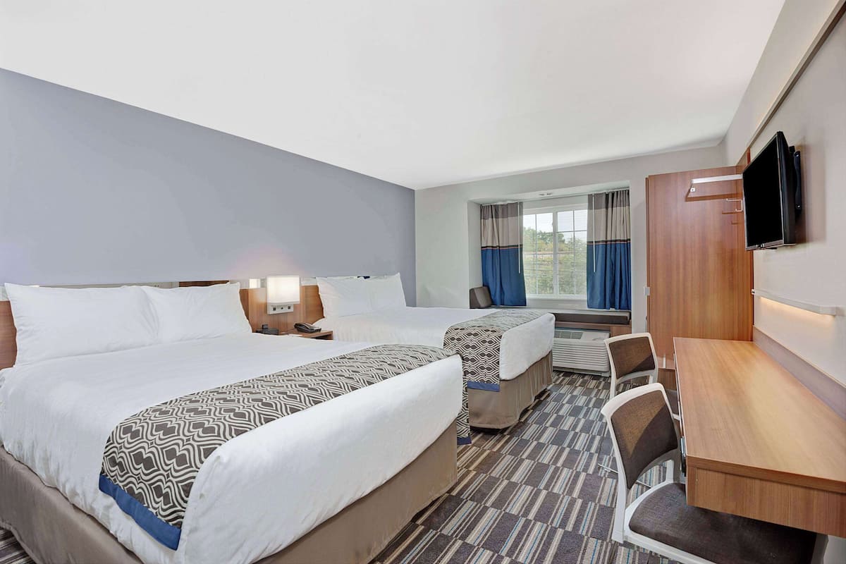 A hotel room with two beds, a desk, chairs, a TV, and a window with curtains.