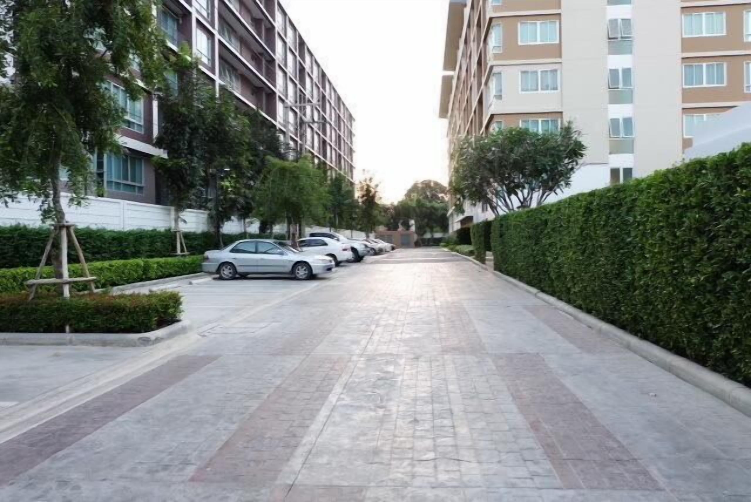 A residential street with parked cars, modern apartment buildings, and well-maintained hedges.