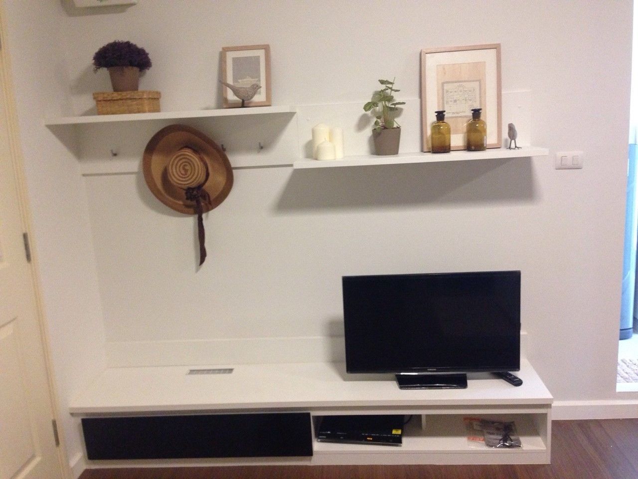 Un salon avec un tél éviseur à écran plat posé sur un meuble blanc, des étagères ornées d’objets décoratifs et une plante en pot.