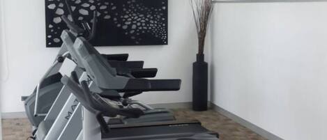 Un gimnasio con cintas de correr y una máquina elíptica, un cuadro abstracto negro en la pared, y una ventana con vistas a la vegetación.