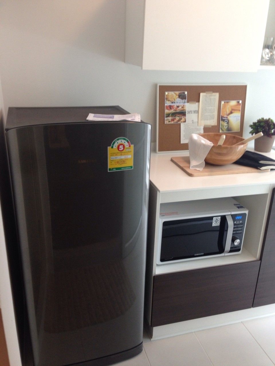 A kitchen with a refrigerator, microwave, and a bulletin board with papers and a plant.