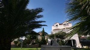 A courtyard with a fountain, palm trees, and a building with a balcony. - Apartments Villa Jadranka - Adults Only (Bol)