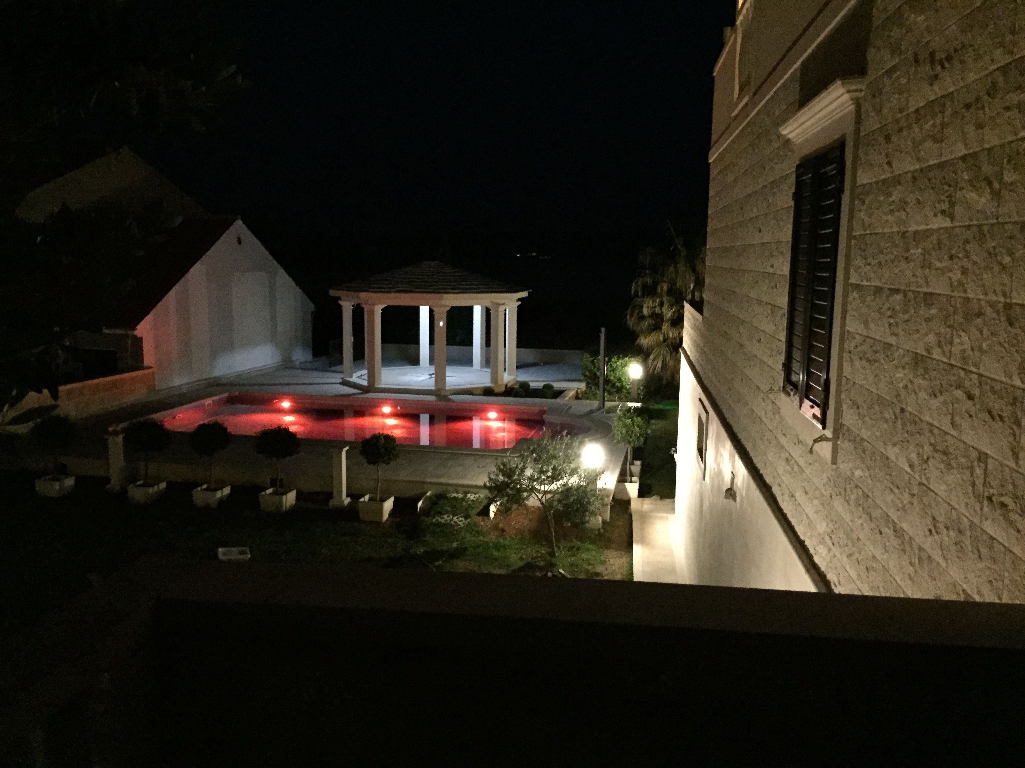 Vue nocturne d’un espace piscine avec un kiosque, entouré d’un bâtiment en pierre doté de volets.