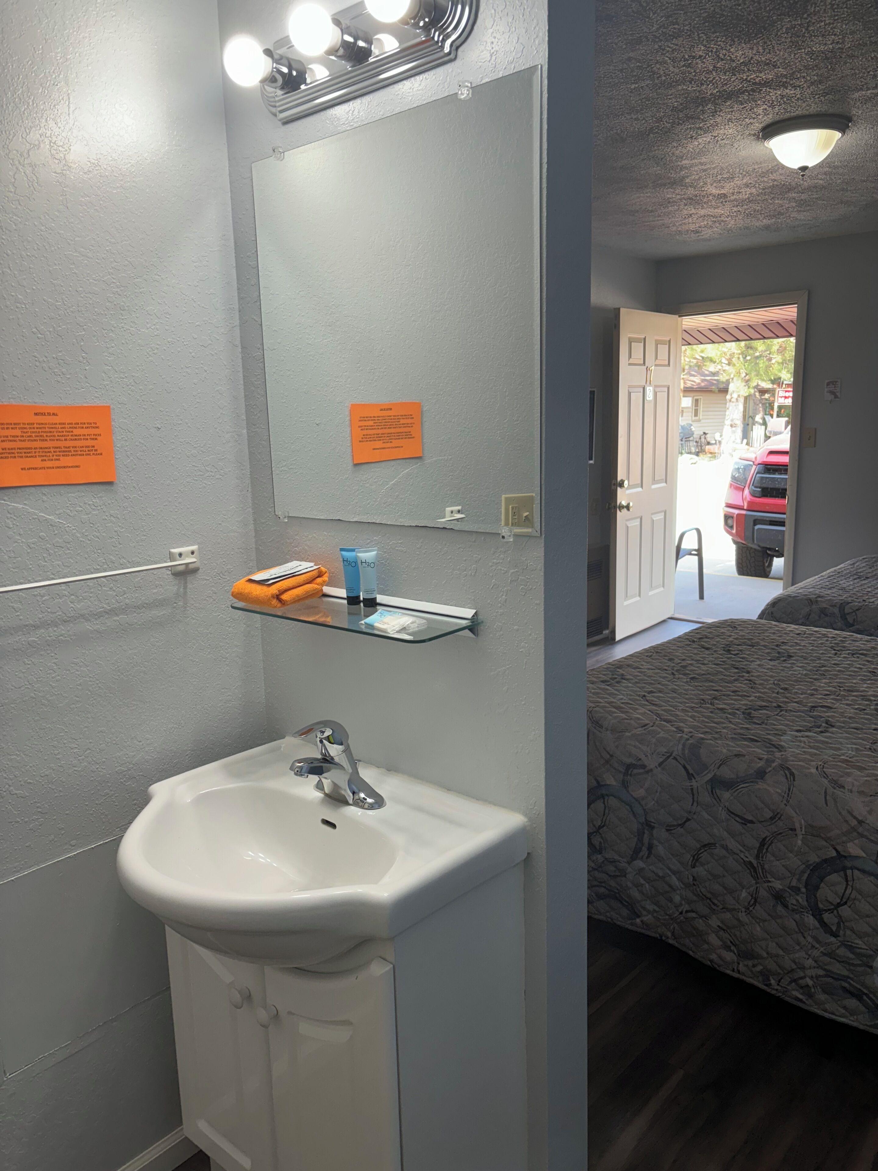 Un baño con lavamanos, espejo y artículos de aseo. En la habitación contigua, hay una cama con cubrecama estampado.