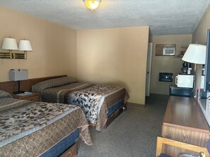 A hotel room with two beds, a desk, a chair, a lamp, a ceiling light, and a door leading to a kitchenette. - Sioux Motel (Murdo)
