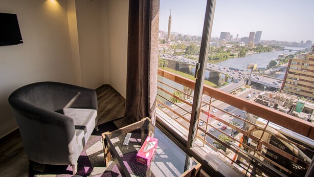 A room with a view of a cityscape, a chair, a small table with a box, and a window with a curtain. - Nile Boutique hotel (Giza)