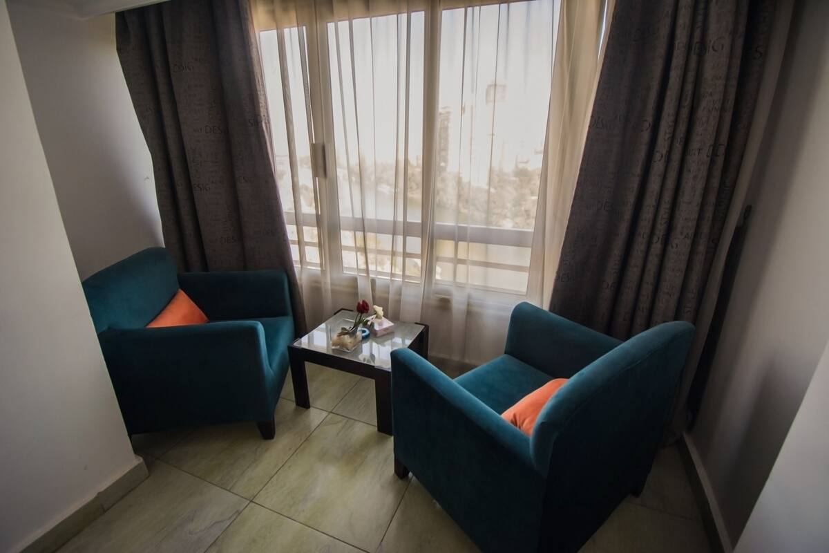 A small room with two teal armchairs, a glass coffee table, and a view through a large window with sheer curtains.