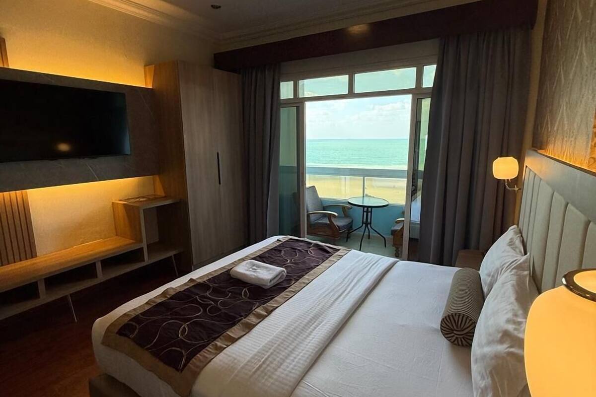 A hotel room with a bed, a TV, a desk, and a view of the beach.