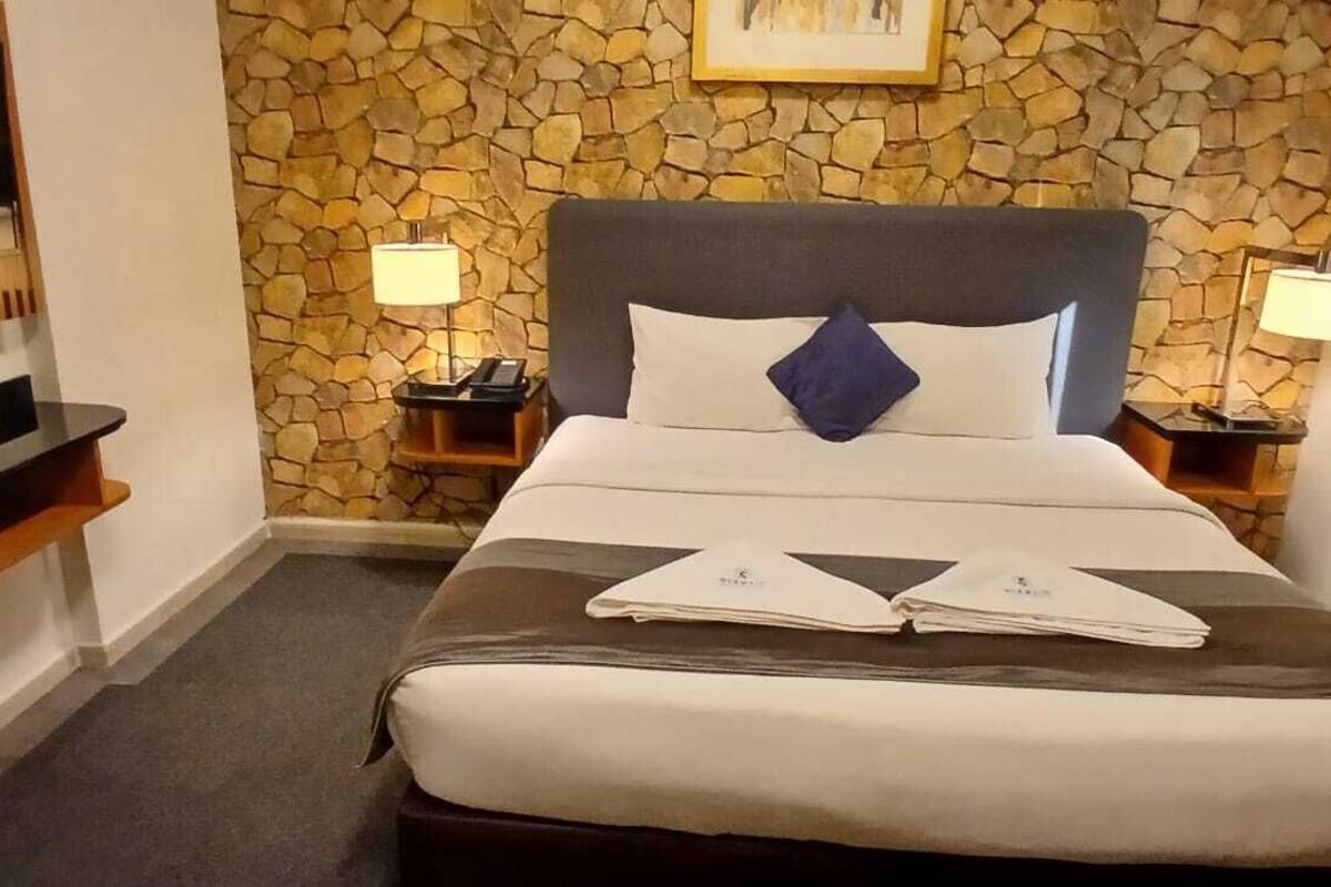 A hotel room with a stone wall, a bed with white and brown bedding, two bedside lamps, a flat-screen TV, and a wooden shelf.