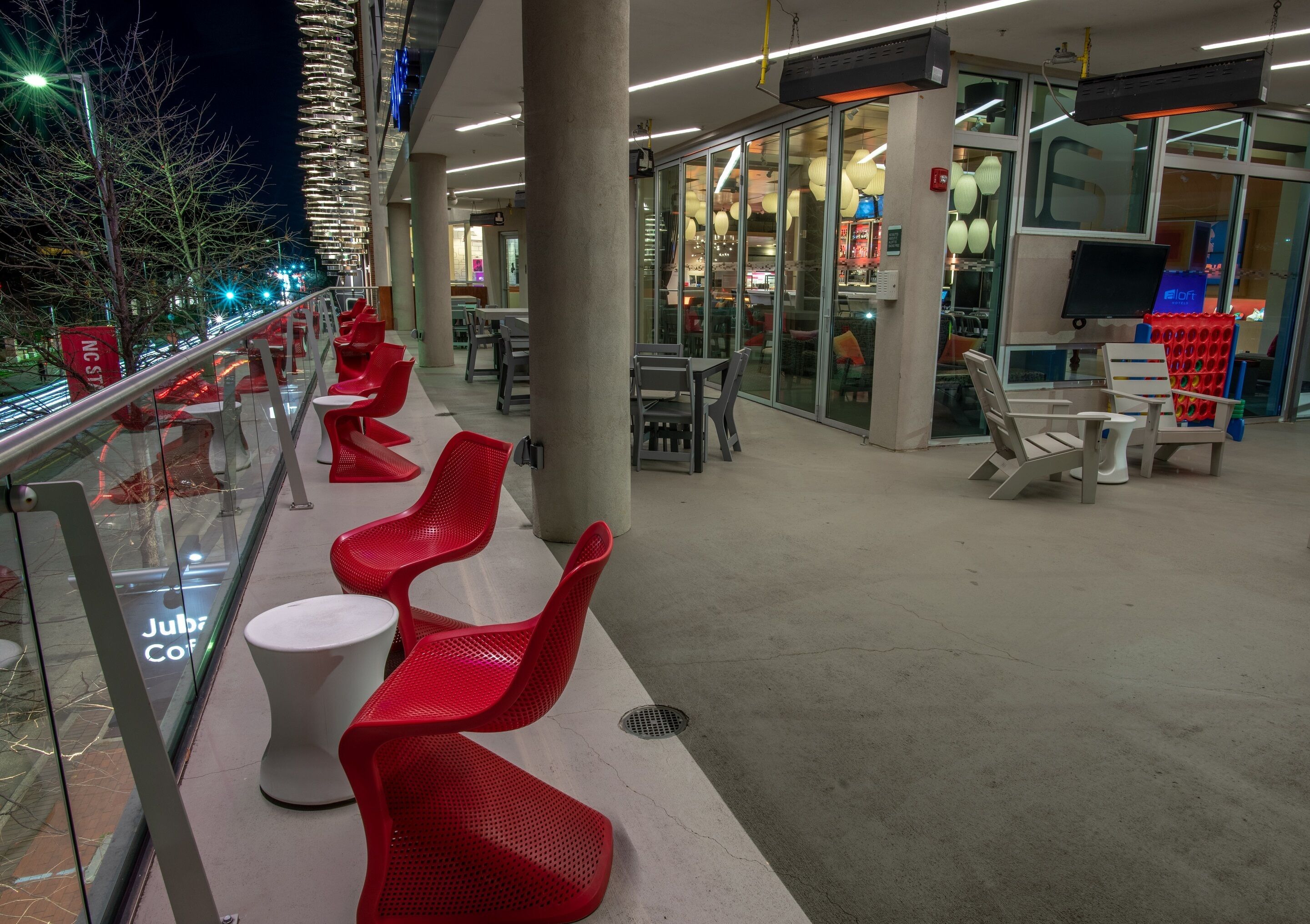 A modern indoor seating area with red chairs and white tables, a glass railing, and a view of the street outside.