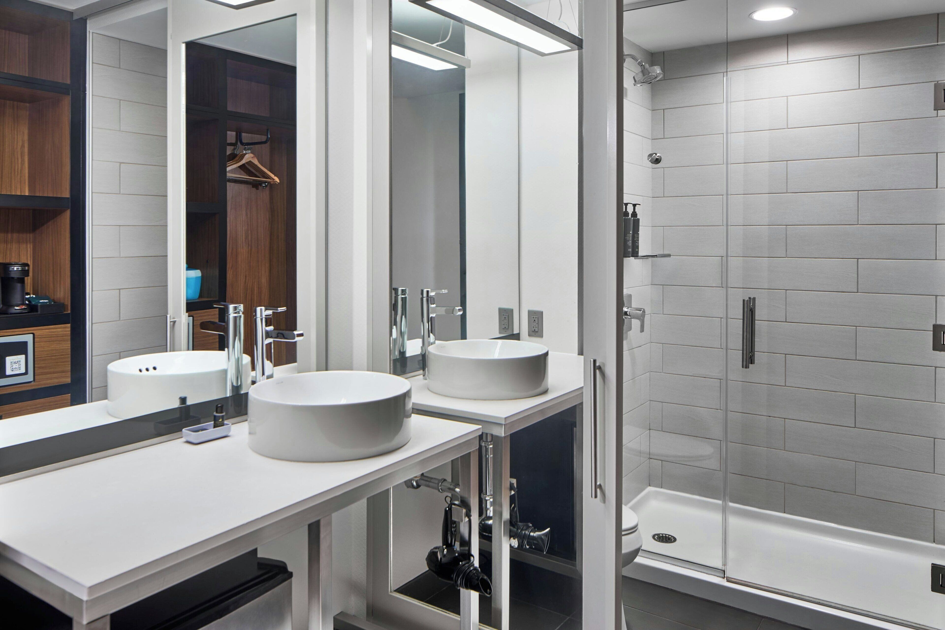 A modern bathroom with two sinks, a large mirror, and a walk-in shower.