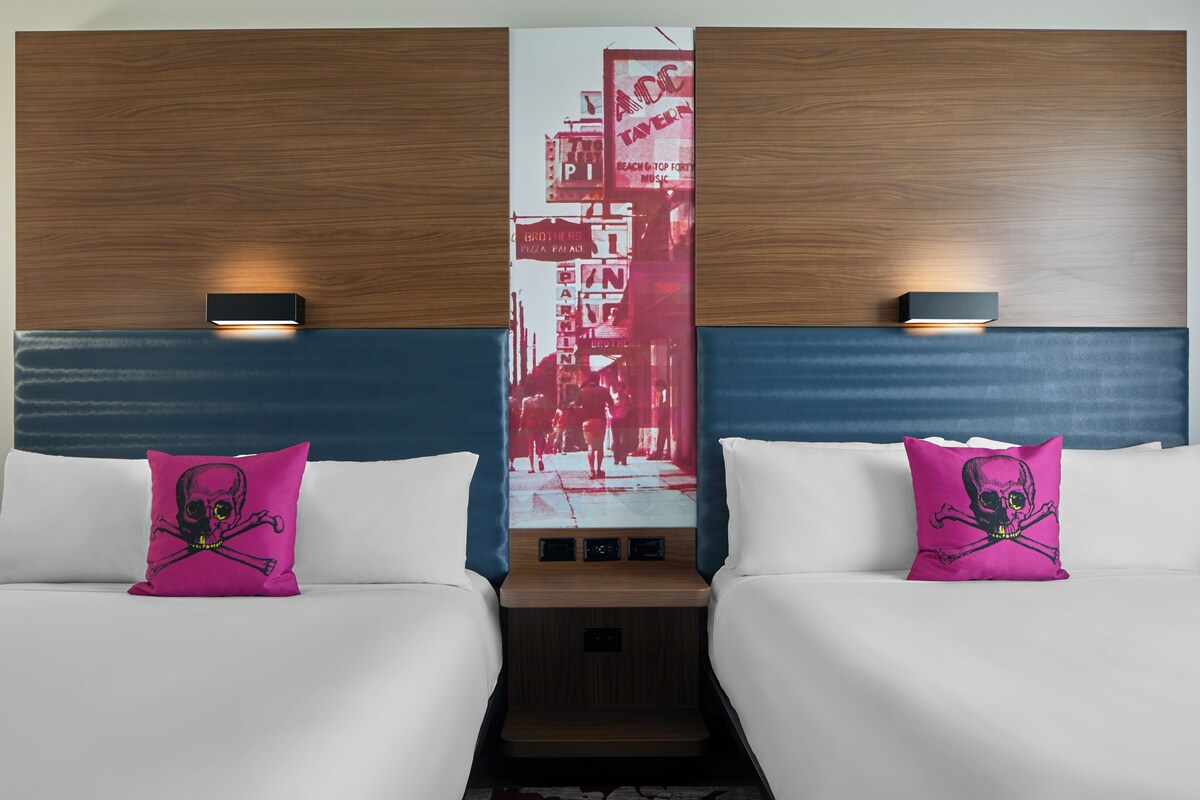 A hotel room with two beds, each with a pink pillow featuring a skull and crossbones design. The room has a wooden headboard and a large cityscape artwork on the wall.