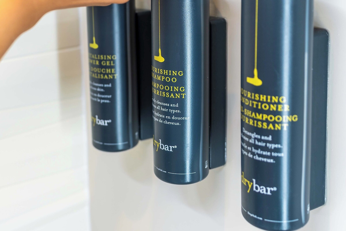 A hand adjusting a black pump dispenser on a wall-mounted shelf with three bottles of hair care products.