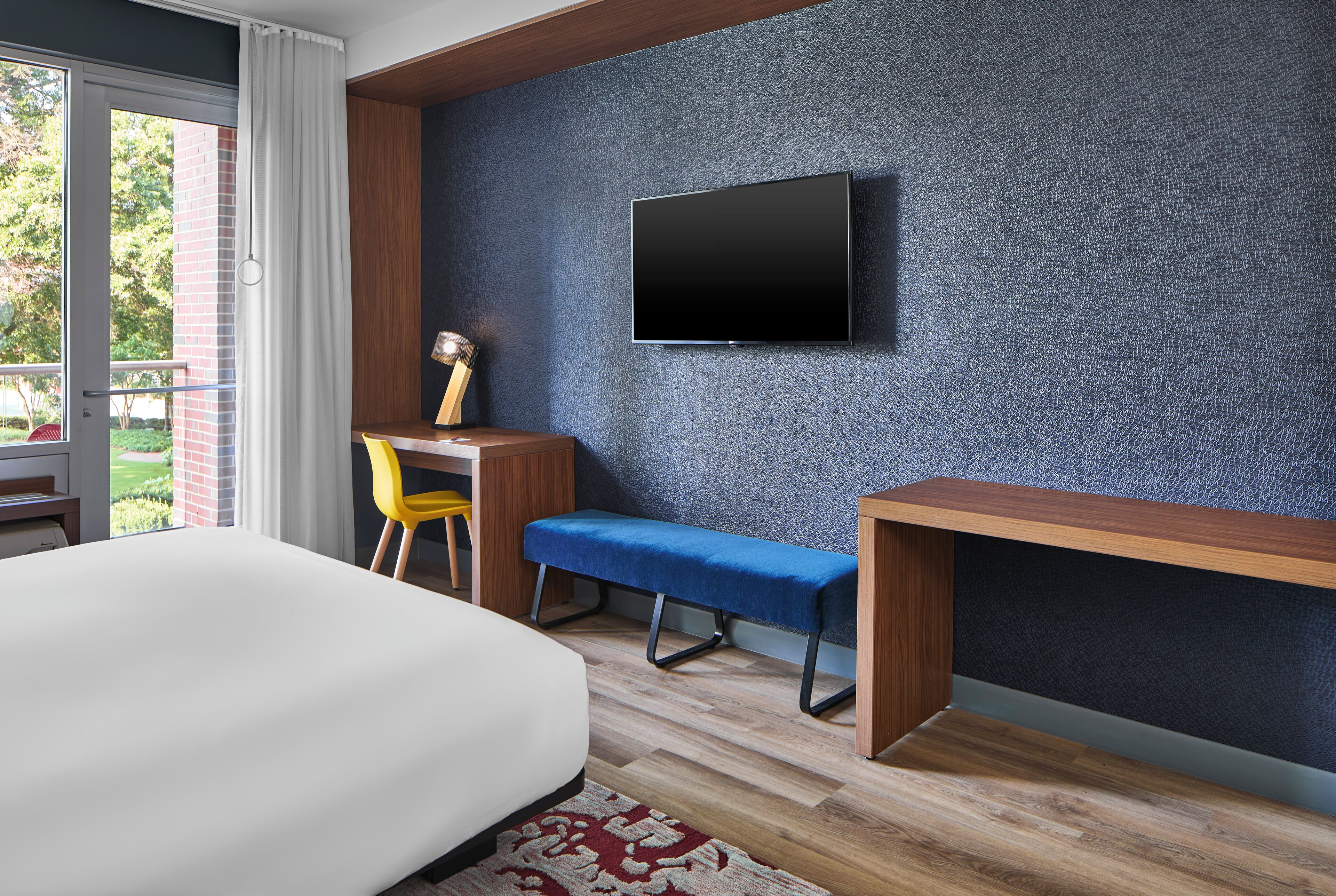 Une chambre d’hôtel moderne, dotée d’un grand lit, d’un bureau avec une chaise, d’un banc et d’un téléviseur fixé au mur.