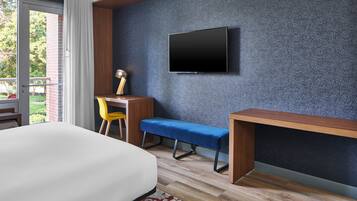 Quarto de hotel moderno com uma cama grande, uma escrivaninha com cadeira, um banco e uma TV montada na parede.