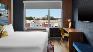 A hotel room with a large bed, a desk, a chair, a TV, and a window offering a city view.