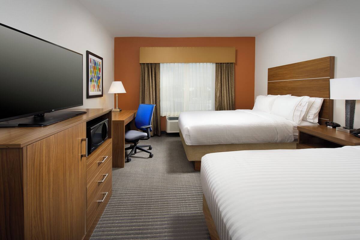 A hotel room with a large flat-screen TV, a desk with a lamp, two beds, a chair, and a window with curtains.