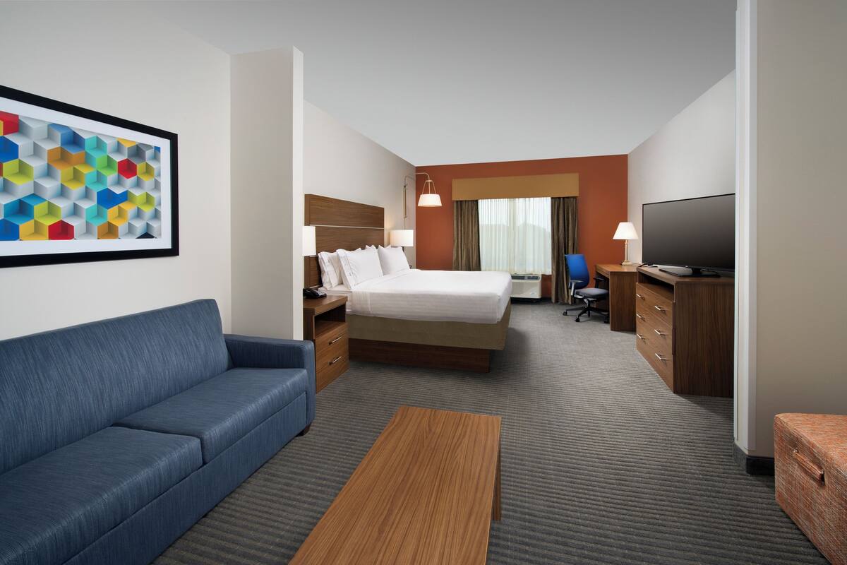A hotel room with a bed, a blue sofa, a wooden coffee table, a TV, and a desk with drawers.