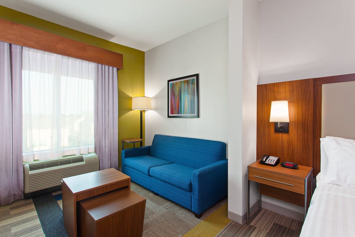 A hotel room with a blue sofa, a wooden nightstand with a lamp, a bed, and a window with curtains.