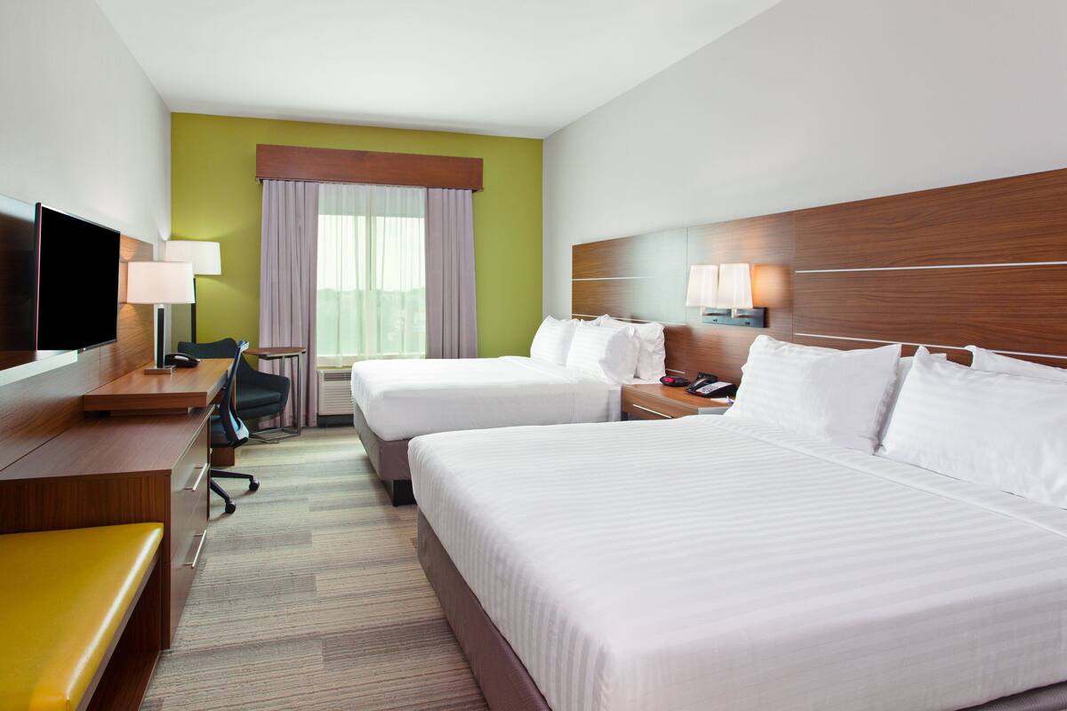 A hotel room with two beds, a desk with a TV, a chair, a lamp, and a window with curtains.