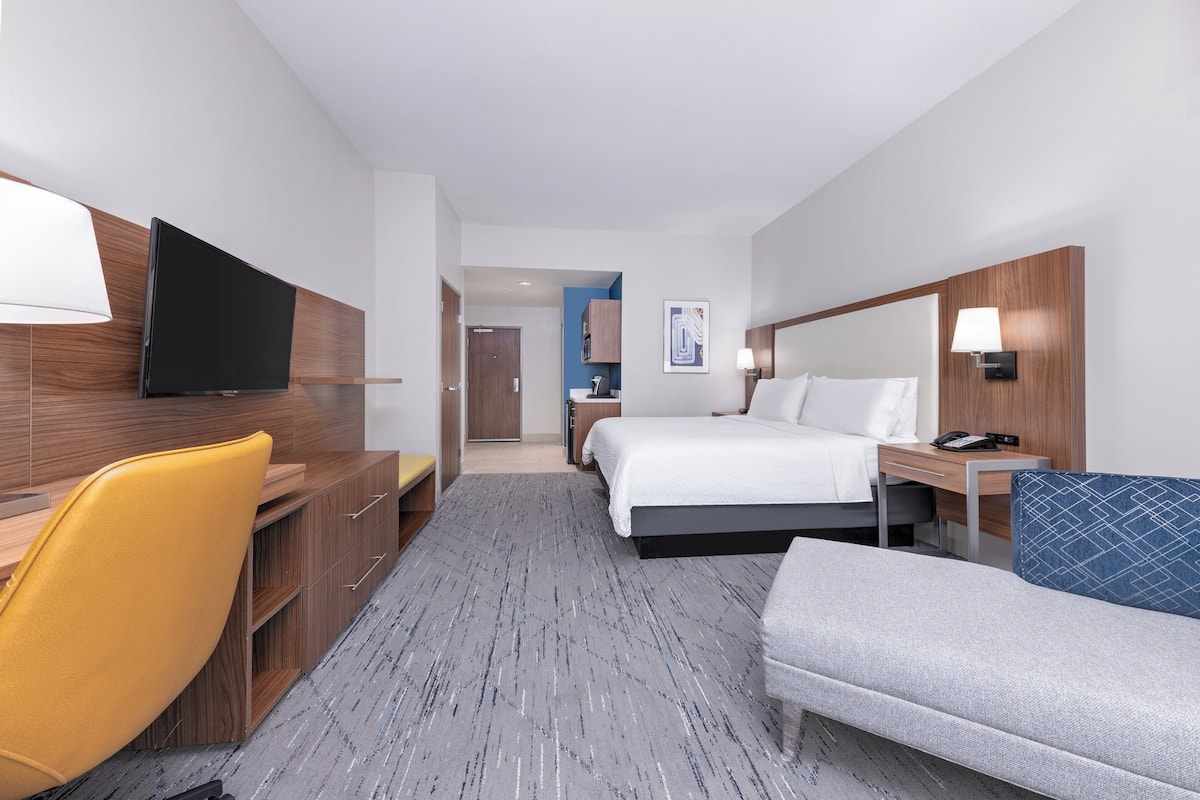 A hotel room with a bed, a desk with a chair, a TV, a bedside table with a lamp, and a small seating area.