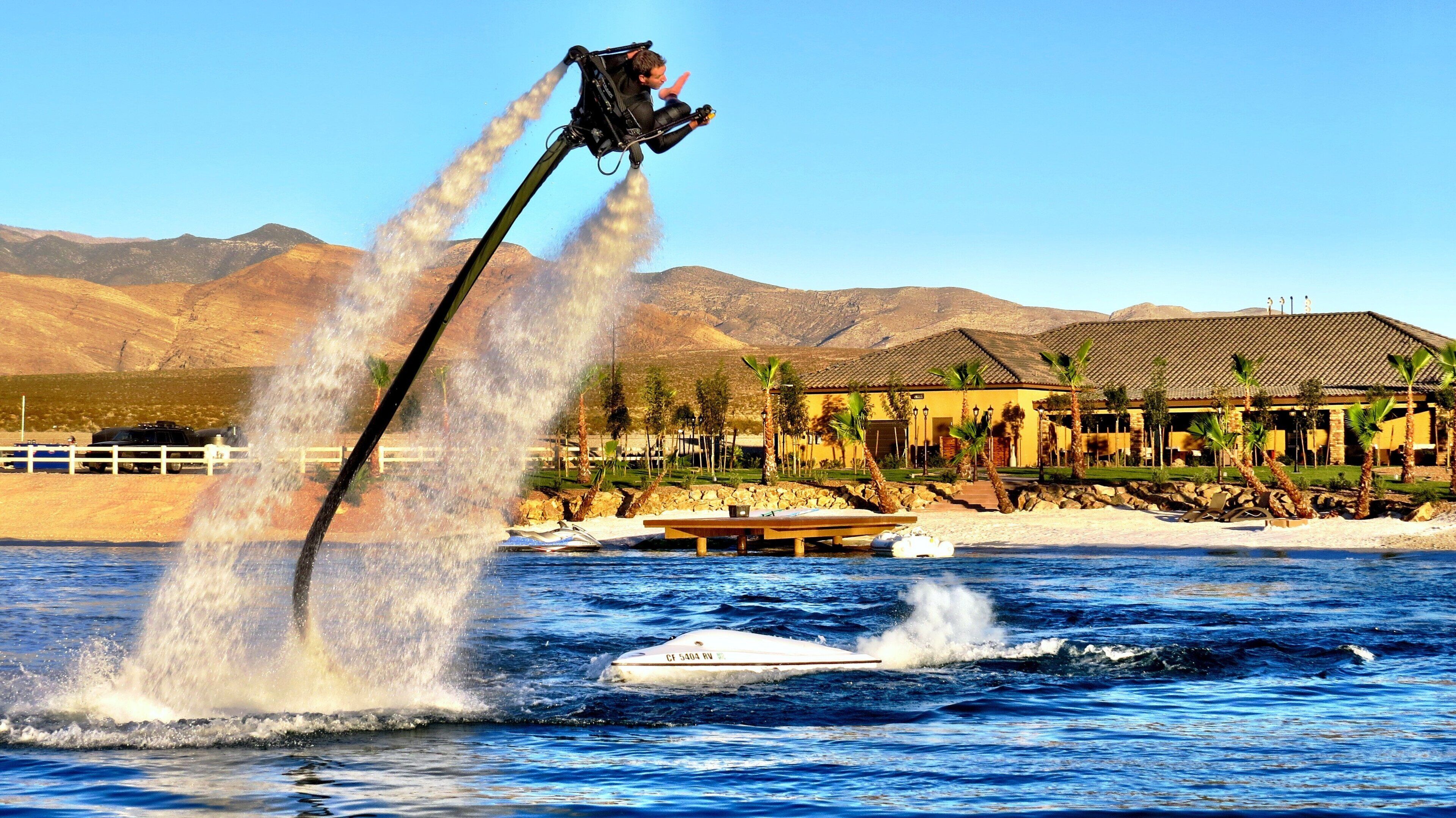 A water jet ski in motion with a person on board, a boat, and a building with a tiled roof in the background.