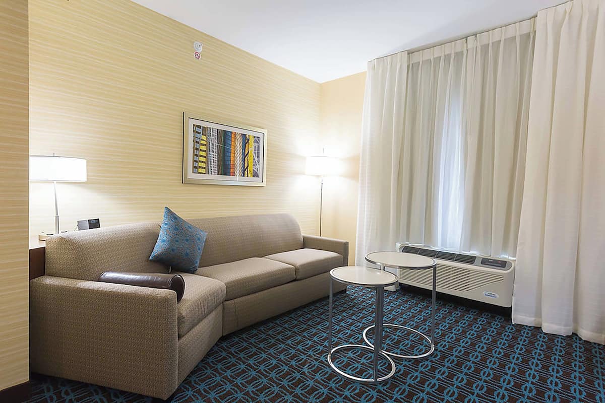 A hotel room with a sofa, a small table, a lamp, a heater, and a window with curtains.