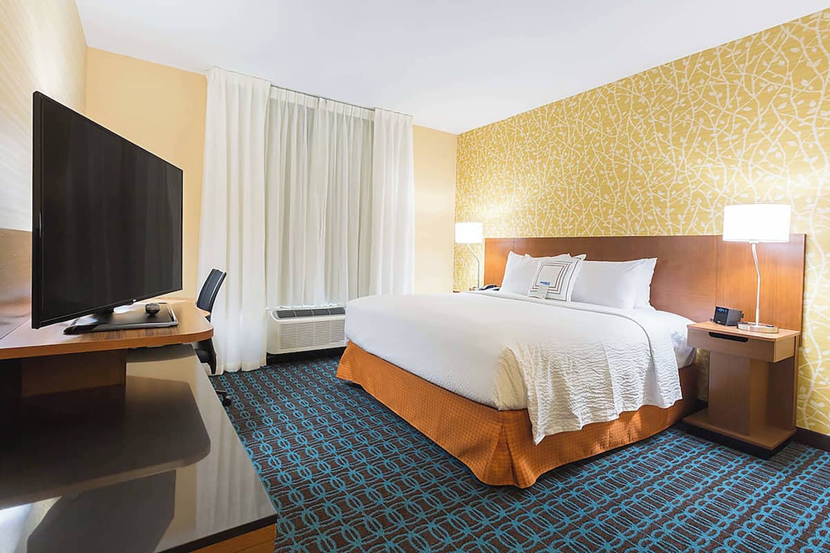 A hotel room with a bed, a desk with a flat-screen TV, an air conditioning unit, and a patterned carpet.