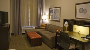 Une chambre d’hôtel équipée d’un canapé, d’un pouf, d’un bureau, d’une chaise, d’une lampe et d’œuvres d’art accrochées aux murs.